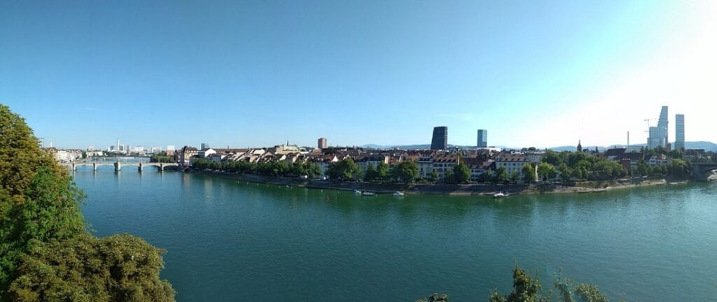 Blick auf den Fluss, der durch Basel fließt und die andere Uferseite bei blauem Himmel.